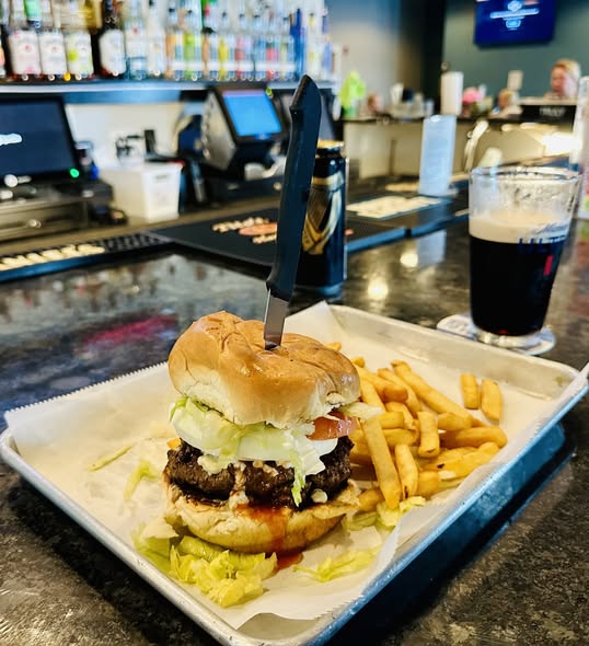 Tall burger with a knife through it, dark beer beside it, bar bottles in background